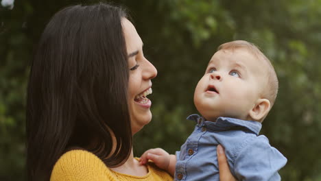 Retrato-De-Una-Madre-Alegre-Sosteniendo-A-Su-Lindo-Hijo-Y-Sonriendo-A-La-Cámara-En-El-Parque