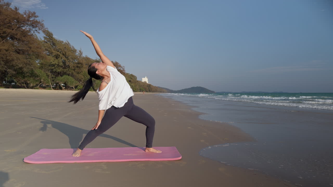 Donna asiatica che pratica la posizione yoga del guerriero inverso su una spiaggia tropicale in Thailandia. Scena di fitness e benessere all'aperto ideale per contenuti di viaggio, salute e stile di vita.