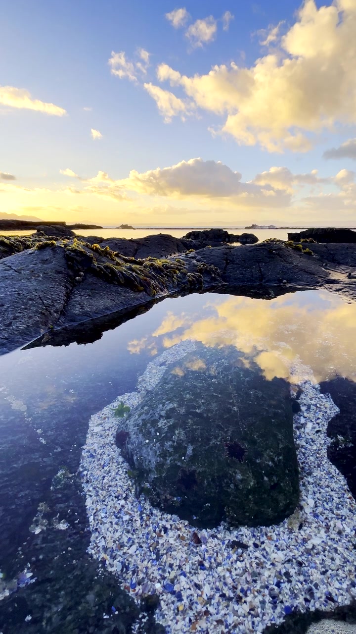 Varmt oplyste solopgangsskyer spejlet i et roligt klippebassin i havet. Vertikal.