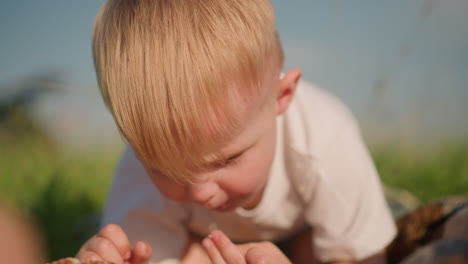a close-up image of a little boy in a white shirt, bent over, deeply focused on eating a piece of bread. the background is blurred, grassy environment that highlights the peaceful outdoor moment