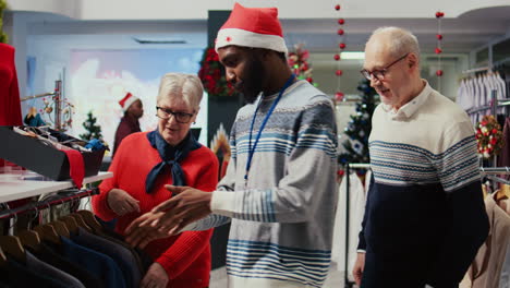 Wife-and-husband-in-Christmas-decorated-shopping-mall-clothing-store,-being-assisted-by-african-american-employee-with-finding-perfect-outfit-during-winter-holiday-season-promotional-sales