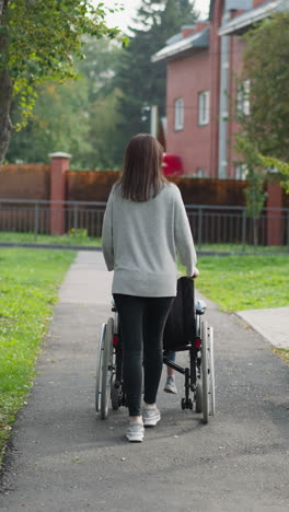 mujer camina por el pavimento de asfalto en el parque empujando a un niño sentado en silla de ruedas. madre e hija con parálisis disfrutan del aire fresco vista trasera