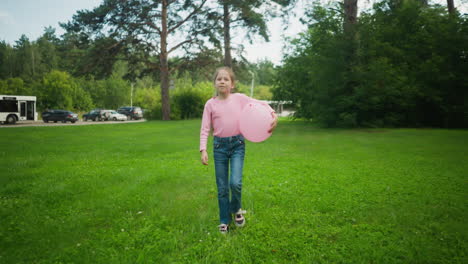 little kid jumping happily while holding pink balloon under arm, hair swaying behind as she walks across green field with parked cars and moving traffic visible in background on sunny outdoor day