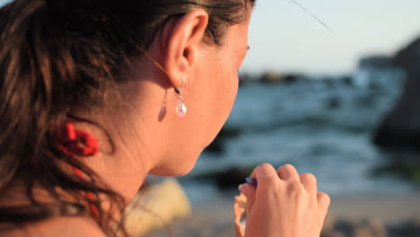 Toma-Lenta-De-Una-Mujer-Hermosa-Con-Un-Aspecto-Natural-Comiendo-Con-Una-Cuchara-Un-Helado-En-La-Playa-Mientras-Las-Olas-Se-Rompen-Y-El-Sol-Ilumina-Su-Rostro-Y-Disfruta-De-La-Vista-Al-Mar