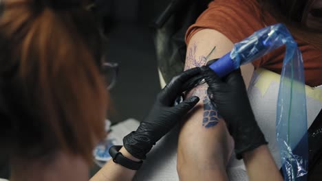 The-girl---the-master-of-tattoos.-Abstract-pattern-tattoo.-The-tattoo-artist-at-work.-A-red-headed-girl-holding-a-tattoo-machine-and-applied-in-a-pattern-on-the-skin-of-her-female-client,-checking-the-sketch-picture.-Footage-from-the-shoulder