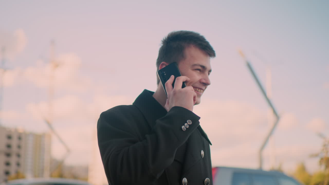 검은 코트를 입은 프리랜서가 웃으며 흥분하여 전화로 이야기하면서 현대적인 건물, 부드러운 빛 및 차량을 특징으로하는 활기찬 도시 지역에서 몸짓을 하고 있습니다.