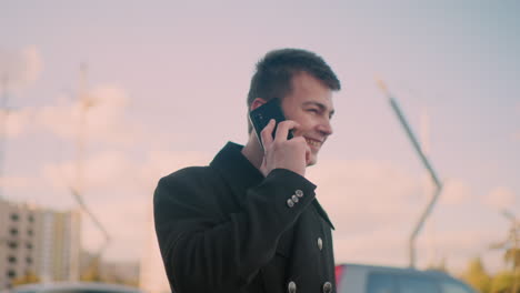 vista lateral de un trabajador independiente con abrigo negro sonriendo y hablando por teléfono con emoción mientras hace gestos en una vibrante área urbana con edificios modernos, luz solar suave y vehículos