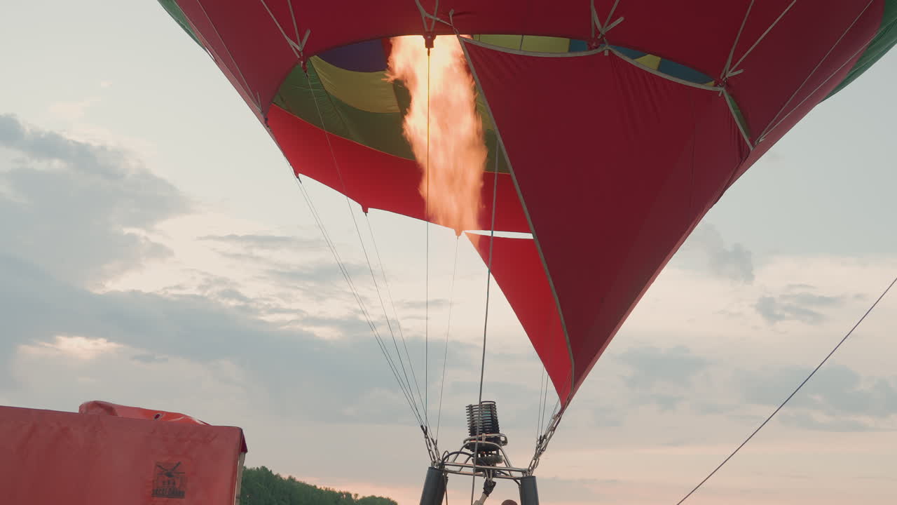 passenger stepping into wicker basket gripping burner controls and tether ropes on grassy sunset field while crew adjusts flame preparing hot air balloon takeoff under open sky for aerial adventure