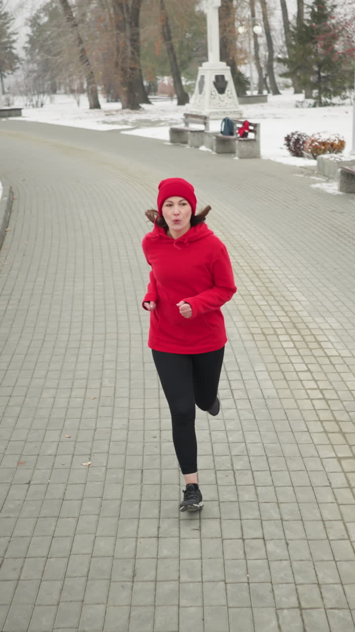 back view of woman jogging along snowy park pathway in red hoodie and black leggings by benches bushes iron railing lamp posts and distant cross monument conveying cold fitness motion