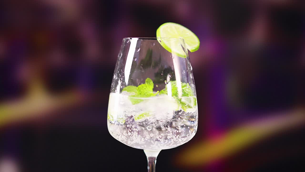 A mojito cocktail with lime and mint in a glass, set against a blurred, colorful background