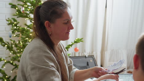side view of woman smiling while carving and cutting paper using scissors boy sitting across watching warmly christmas tree with glowing lights blurred in background cozy holiday atmosphere