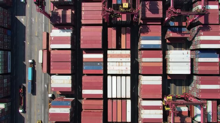 Large shipping containers are seen stacked tightly together at a bustling port. Cranes are actively loading and unloading cargo, showcasing efficient logistics in action.