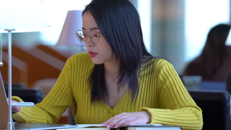 portrait of happy joyful pretty female student busy hard working sitting against background of bookshelves in university library holding laptop and backpack looking to the side, smiling pleasant