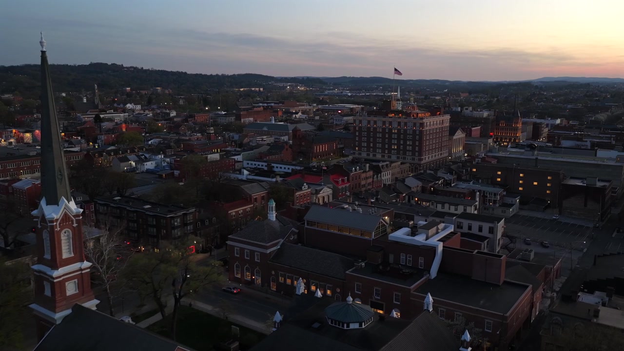 Sorvolo aereo sopra la storica York City, Pennsylvania, al tramonto con la torre della chiesa, i tetti del centro, auto in movimento, la bandiera americana, edifici classici e una calda atmosfera dello skyline serale.