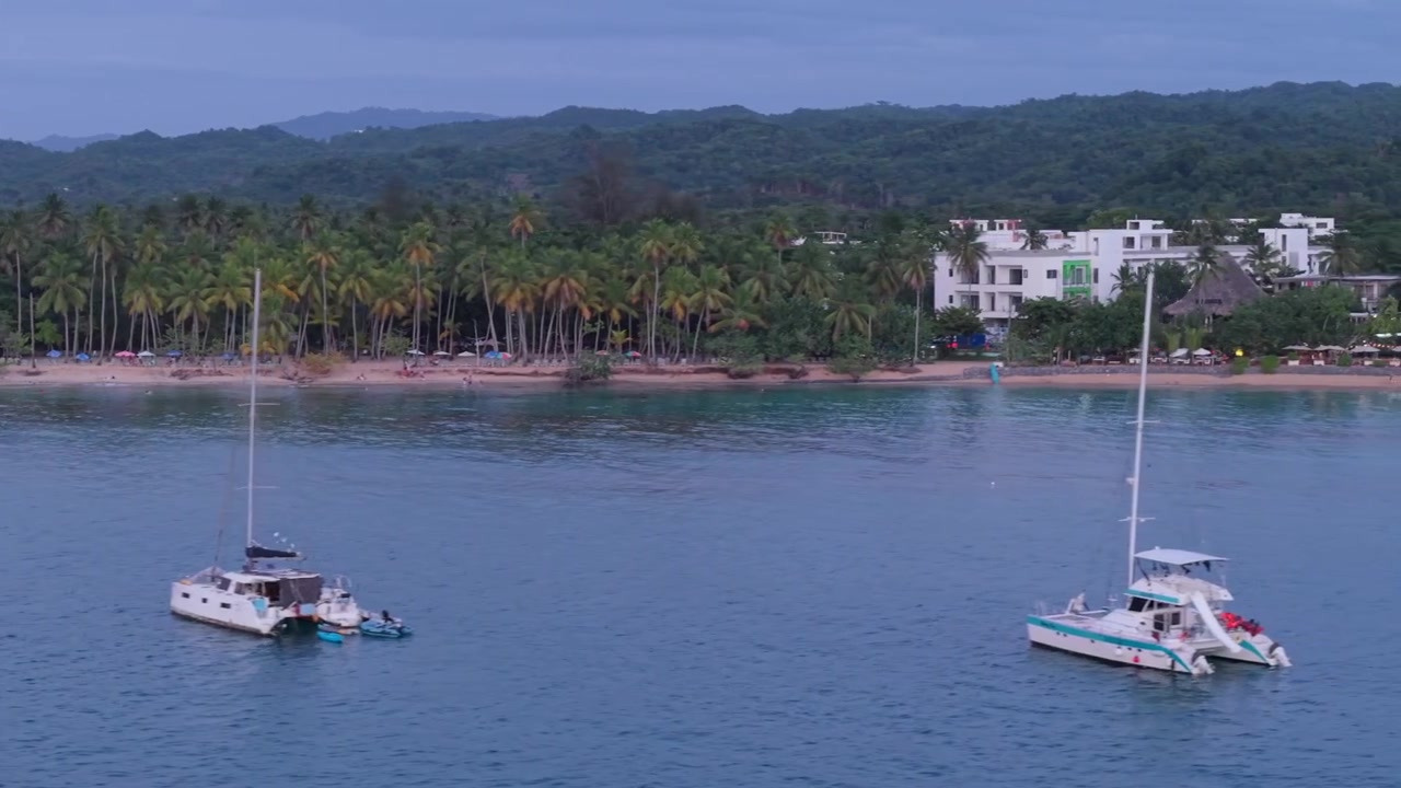 Ripresa aerea con drone in panning che rivela barche a vela e catamarani ancorati in una baia calma, con una spiaggia tropicale orlata di palme ed edifici di resort costieri sullo sfondo.