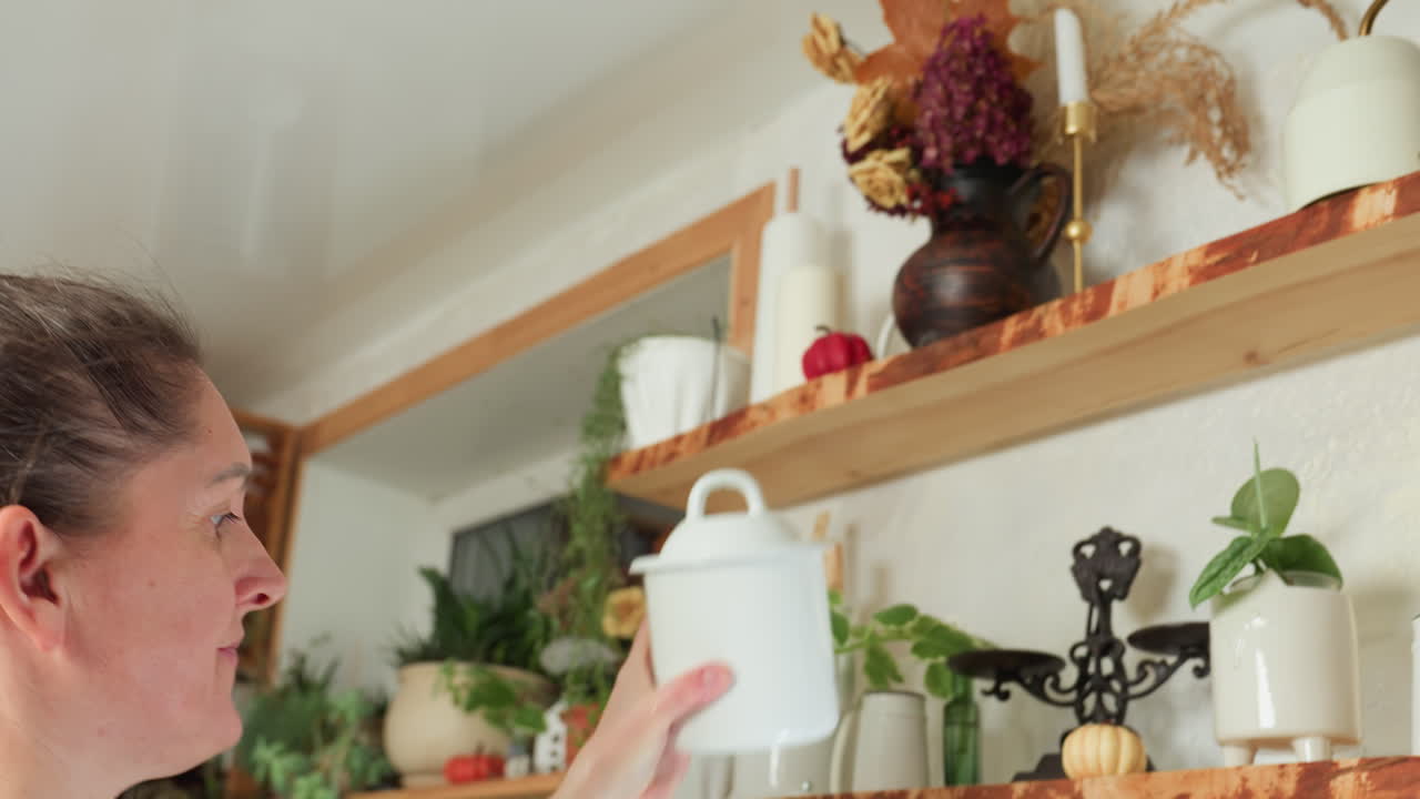 Woman in green dress reaches for ceramic container on wooden shelf in cozy rustic kitchen, preparing to use contents for cooking as natural light brightens space with floral decor elements