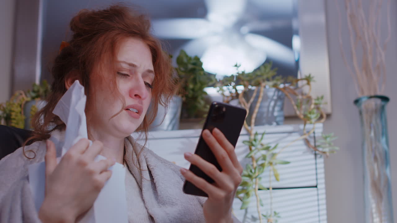 젊은 백인 여성이 집에서 병에 걸려 휴대폰을 쳐다보고 조직 상자를 들고 있습니다.