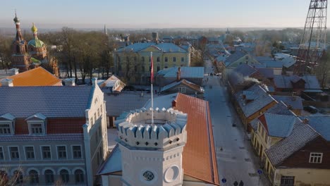 Vista-Aérea-Del-Casco-Antiguo-De-Kuldiga,-Casas-Con-Tejas-Rojas,-Torre-Con-Bandera-De-Kuldiga,-Día-Soleado-De-Invierno,-Destino-De-Viaje,-Amplio-Disparo-En-órbita-De-Drones