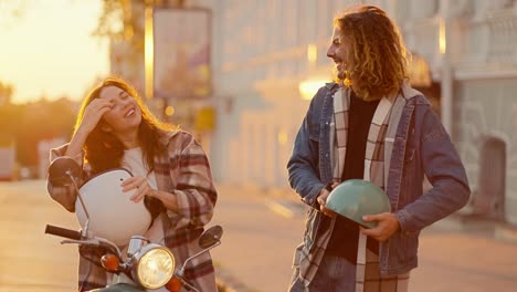 Happy-couple-on-a-golden-sunrise,-a-brunette-girl-sits-on-a-moped-in-a-plaid-shirt-near-her,-her-boyfriend-with-long-curly-hair-in-a-denim-jacket-hugs-her.-They-look-at-each-other-and-rejoice