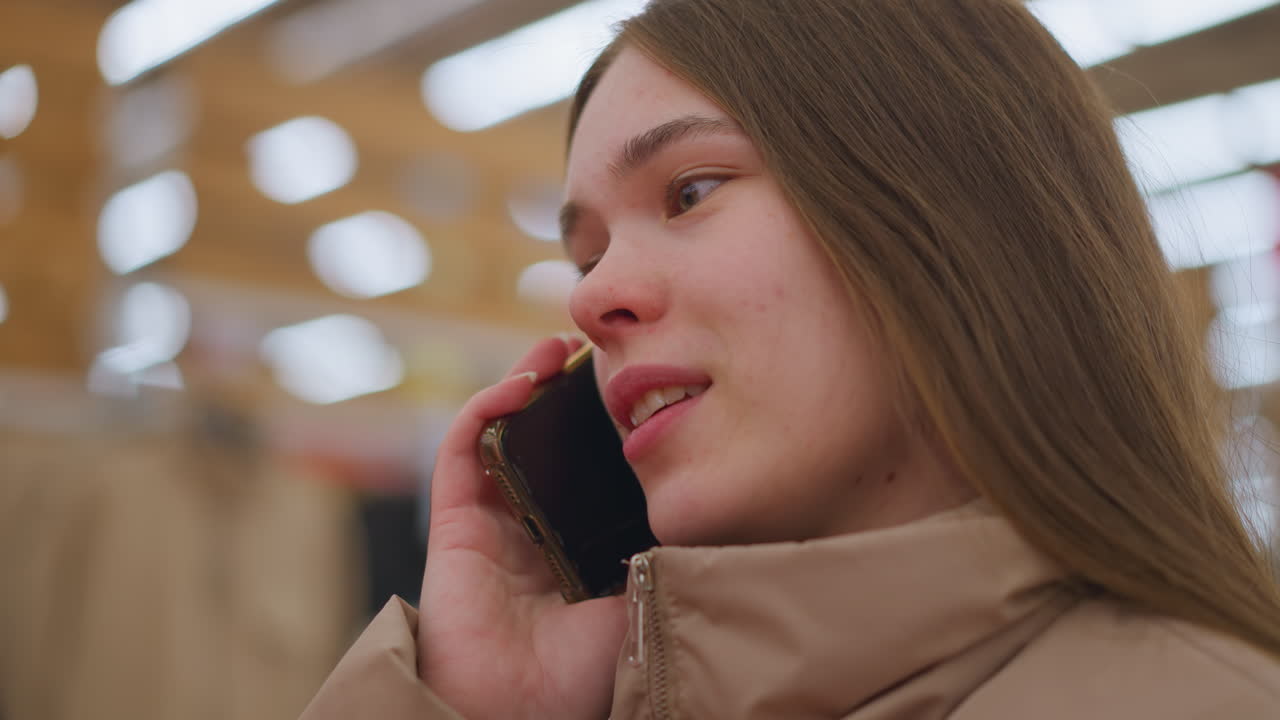 밝은 갈색 재을 입은 소녀가 쇼핑몰에 서서 드폰을 귀에 대고 웃으며 이야기하고 있습니다. 클로즈업은 그녀의 표현적인 얼굴과 대화의 캐주얼한 분위기를 포착합니다.