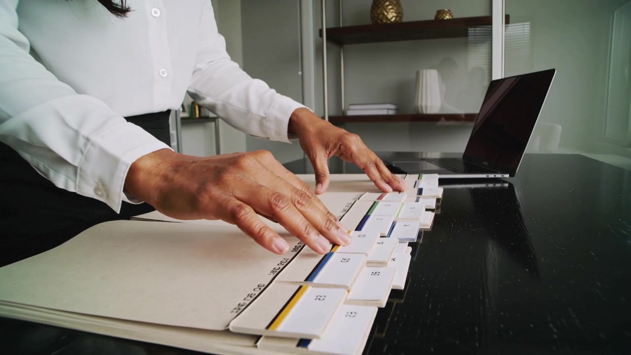 Approaching folder, woman wearing white blouse aligning numbered tabs across home desk for filing