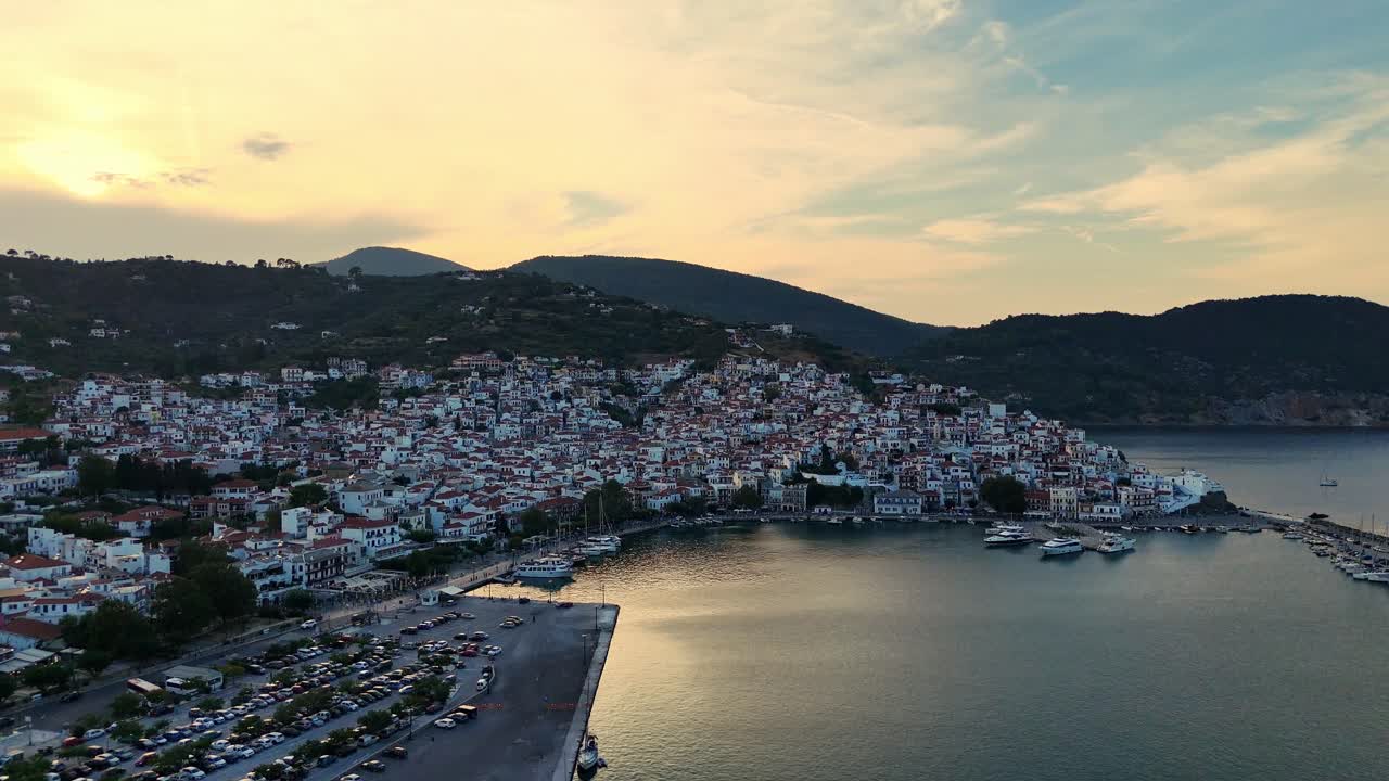 A coastal town at sunset with mountains and boats docked in the harbor, aerial view