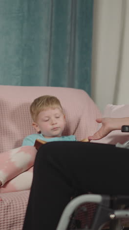 blond son gives book to mother asking to read fairy tale. woman sits in wheelchair spending time with children on pink sofa in living room closeup