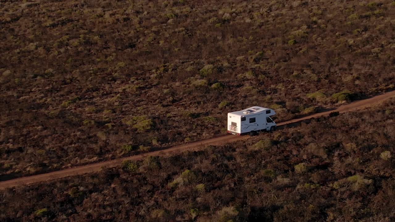 Autocaravana a conduzir por um planalto arenoso acima das Falésias da Arriba Fóssil na Costa da Caparica, Almada, Portugal, atravessando paisagem de falésia fóssil atlântica com vegetação de matagal mediterrânico, acompanhamento por drone