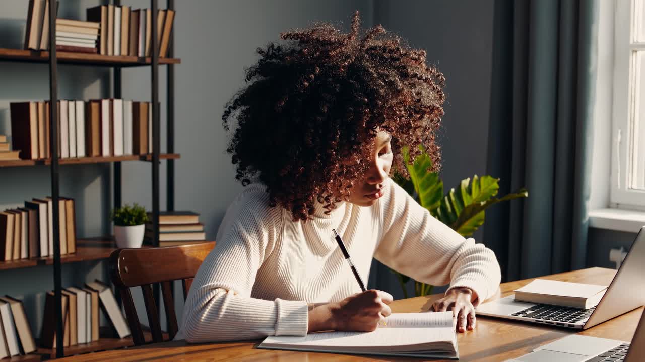 Side angle of a woman writing in a notebook at a desk, with a laptop open, capturing a cozy home