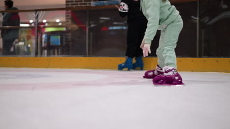 eine nahaufnahme eines kindes in mentengrünen hosen und rosa eislaufen, das auf einer eislaufbahn spielt, das kind wird in der mitte der bewegung gefangen genommen, während es seine eislauffähigkeiten übt