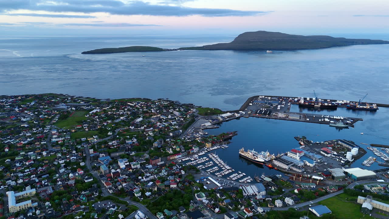 Cinematic aerial view of colorful village houses along a fjord in the Faroe Islands, surrounded by dramatic green mountains, black sand beach, and misty coastal scenery
