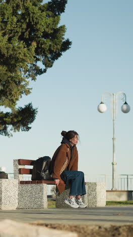 una chica con un abrigo marrón, pantalones vaqueros azules y zapatos blancos se sienta sola en un banco en un parque urbano, su mano descansa en el banco junto a su mochila negra. parece desconsolada y perdida en sus pensamientos