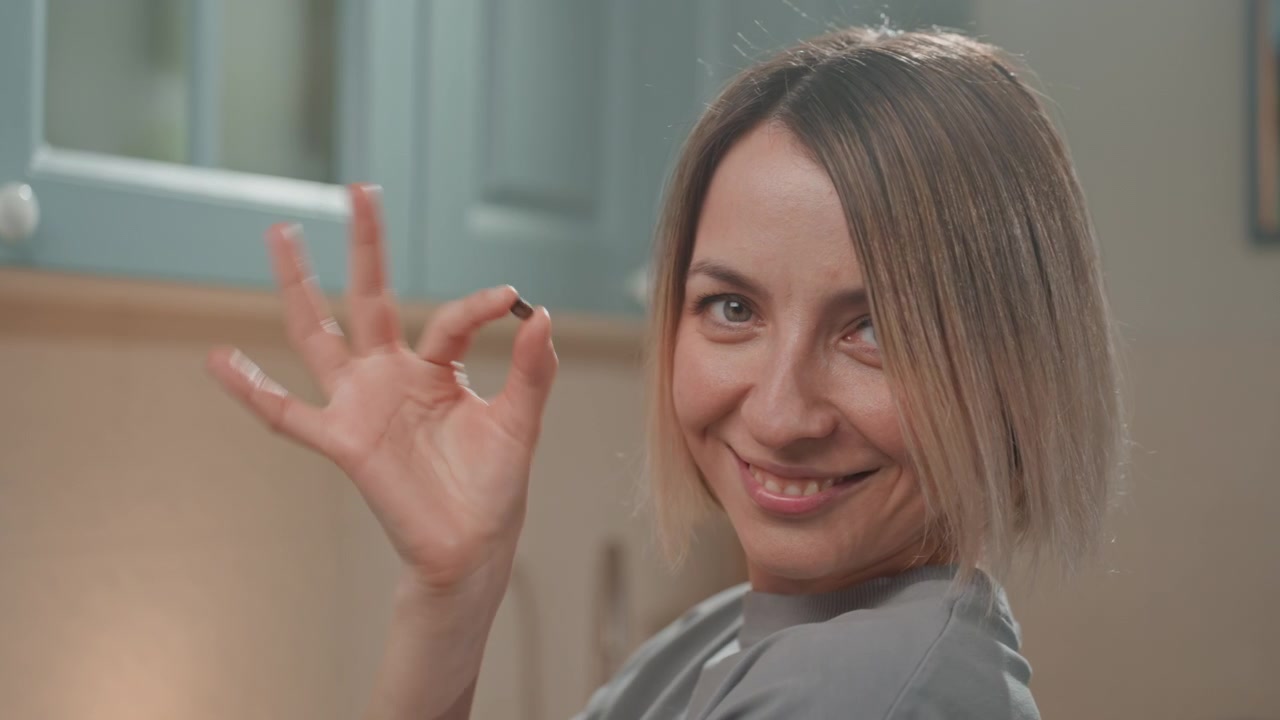 Donna sorridente in cucina fa il gesto dell'ok, espressione giocosa da bacio dello chef, caschetto biondo corto, sfondo di mobili color verde acqua, illuminazione morbida e calda, ritratto ravvicinato di lifestyle di un'amante della musica che si gode il momento.