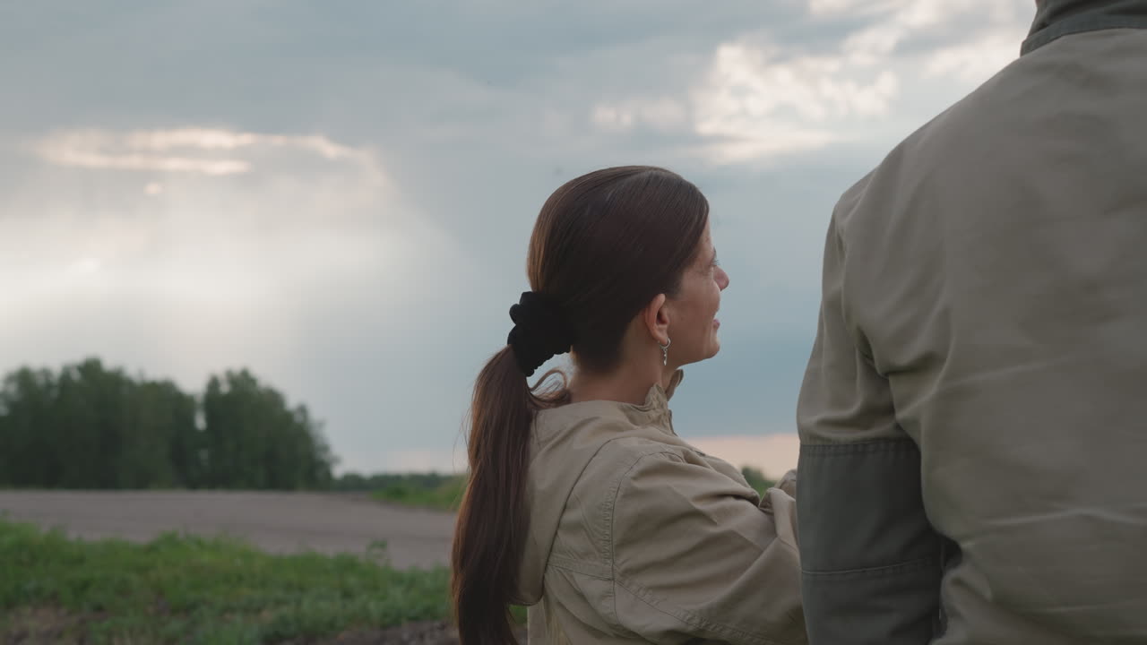 rear view of woman interacting with colleague in open field, smiling and gazing upward at stormy sky with soft sunlight breaking through clouds, hinting teamwork