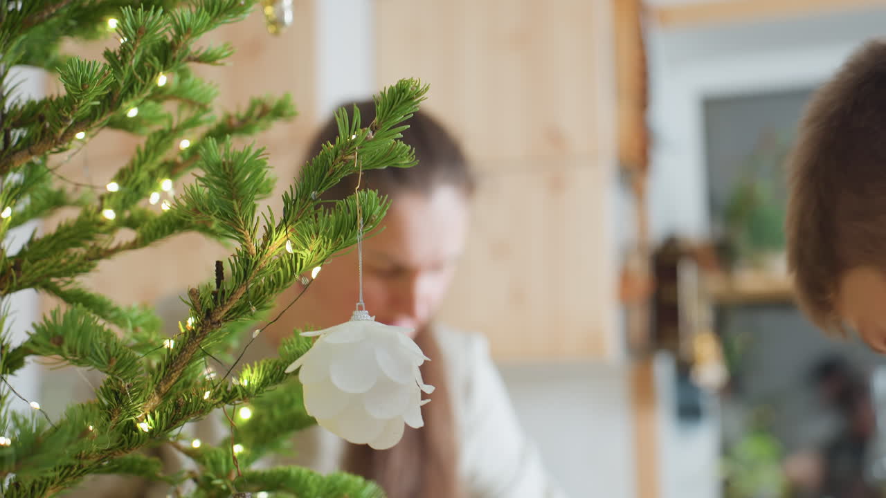 Blurred view of woman fixing ornament on Christmas tree with child assisting beside her, festive green branches decorated with tiny glowing lights, cozy family Christmas preparation