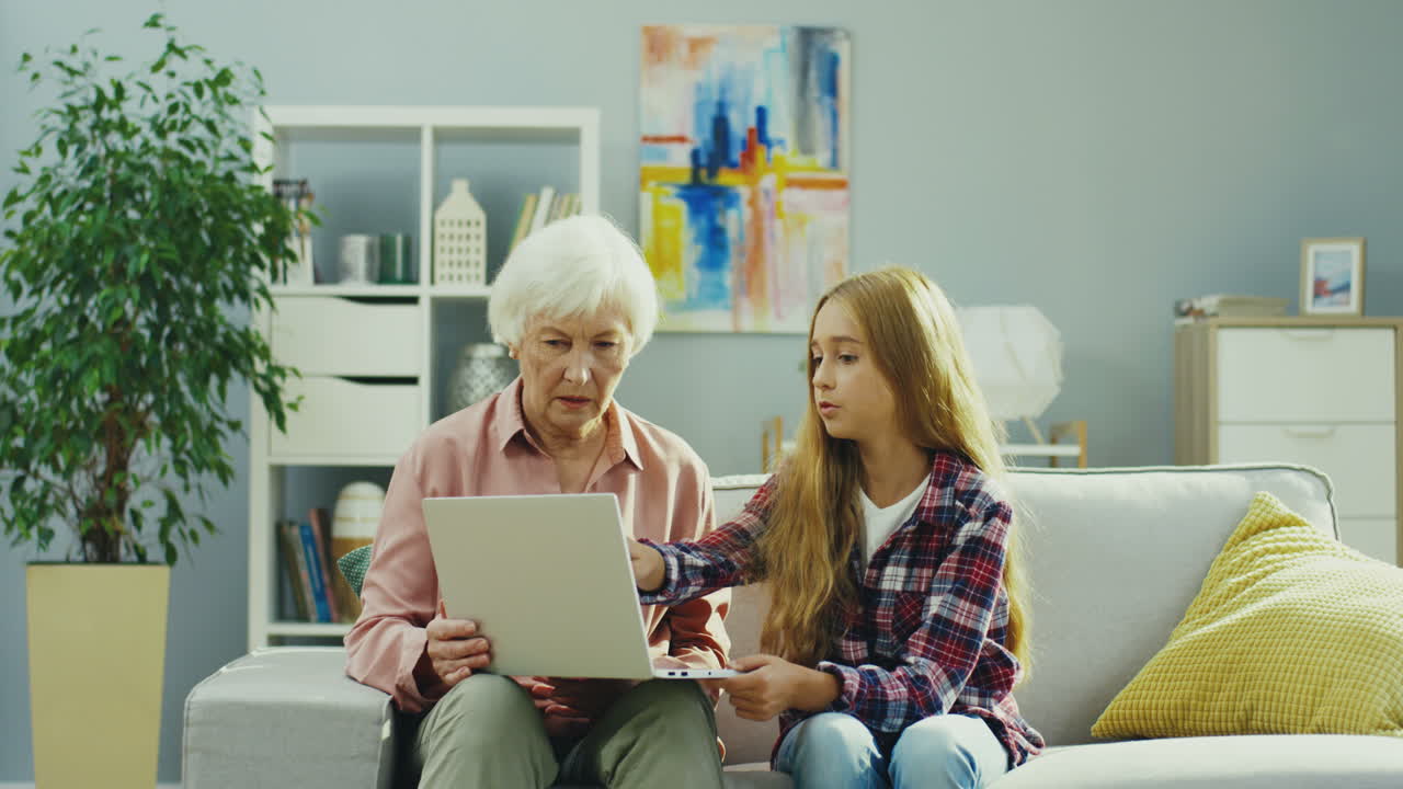 Old Good Looking Woman Sitting On The Couch In The Cozy Livingroom And Telling Something To Her Pretty Teen Granddaughter While Demonstrating Something On The Laptop Screen