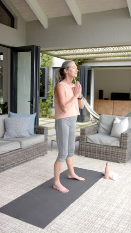 vertical video: practicing yoga, mature woman standing on yoga mat in living room