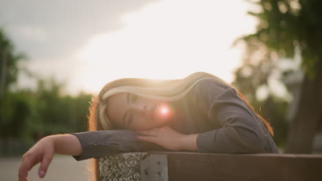 woman seated at edge of bench resting head on arm in serene outdoor park setting with soft sunlight reflecting around her, background includes trees and a building