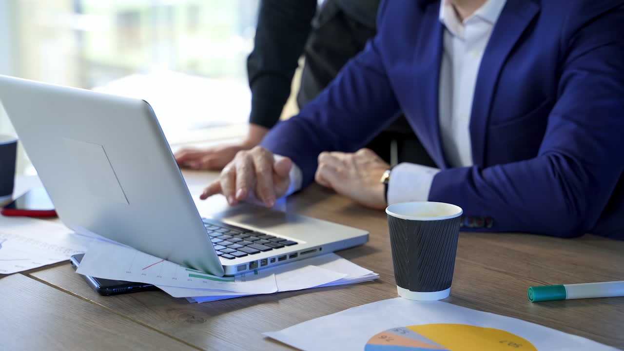 Man working on a laptop at desk. Laptop on office desk and business partners work in it. Male hands on wireless gadget and a plastic cup on table nearby.