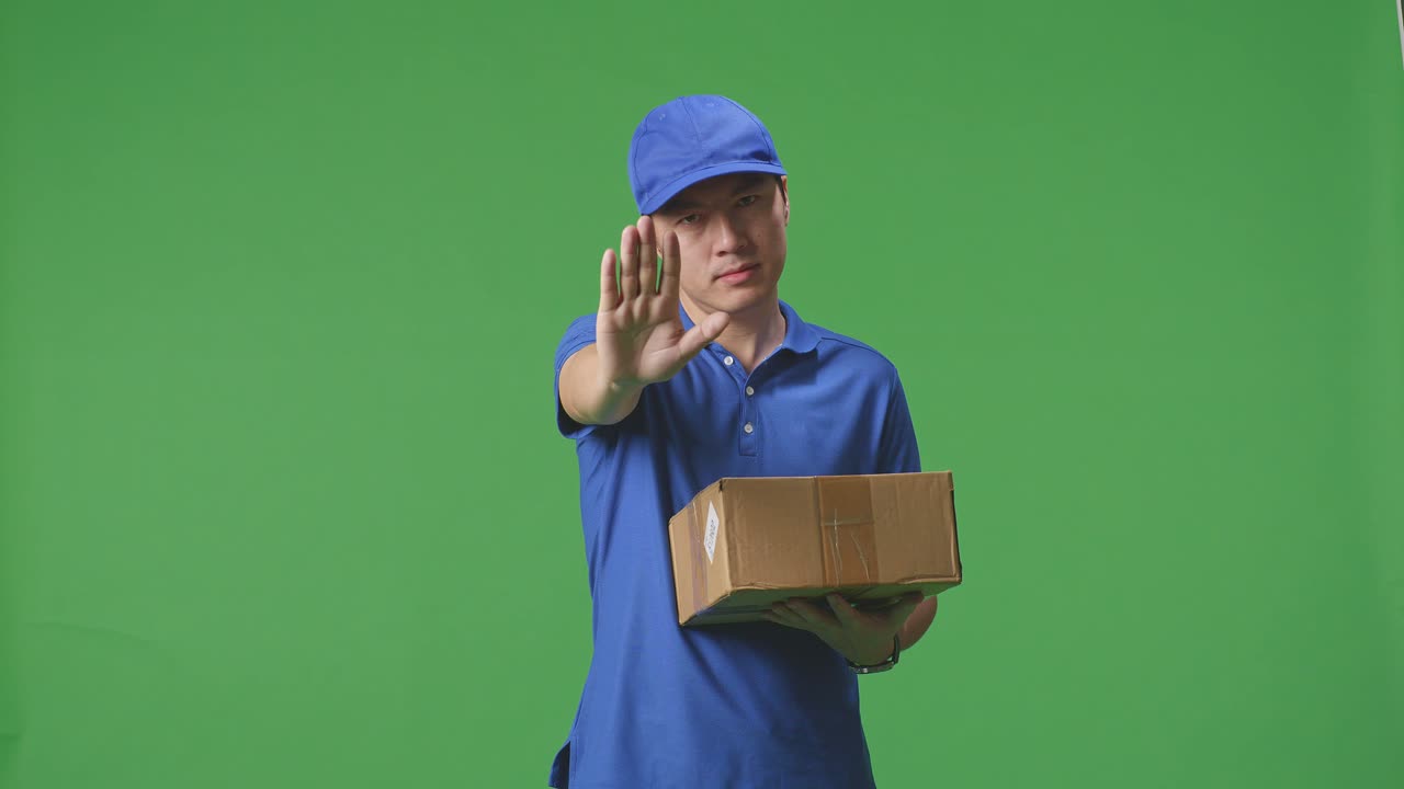 Asian Male Courier In Blue Uniform Making Stop Gesture And Shaking His Head While Delivering A Carton On Green Screen Background In The Studio