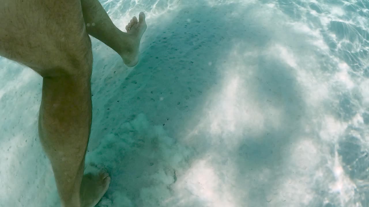 Underwater scene of male legs and feet walking on sandy sea floor with sunlight refractions and reflections in clear transparent ocean water