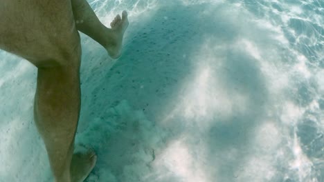 Underwater-scene-of-male-legs-and-feet-walking-on-sandy-sea-floor-with-sunlight-refractions-and-reflections-in-clear-transparent-ocean-water