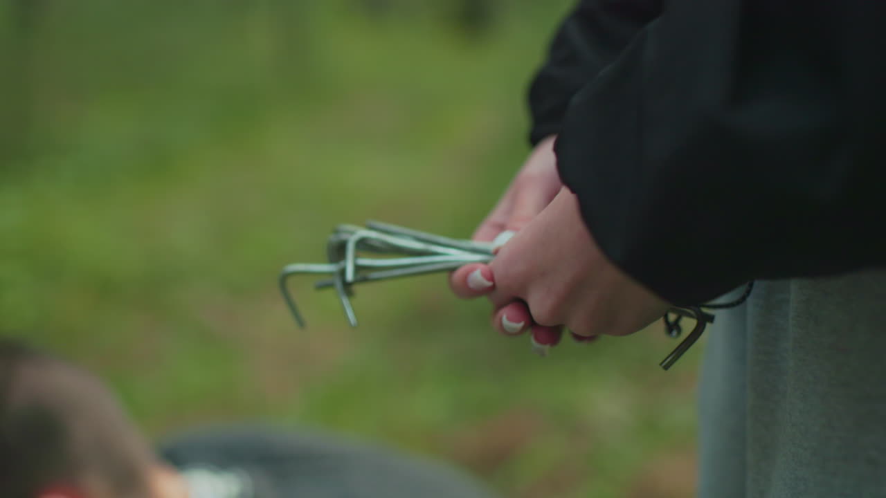 woman gives tent peg to crouching partner holding sturdy wood stick ready to hammer into soft forest ground covered with grass moss and pine needles securing tent stake during couple camping