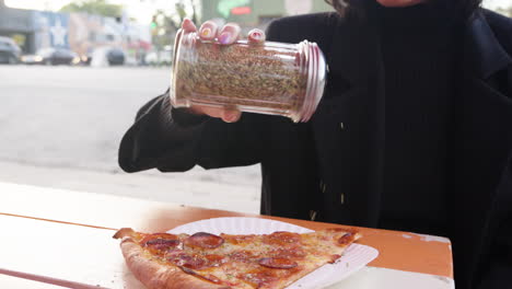 Woman-shakes-oregano-topping-onto-pepperoni-pizza-slice-in-outdoor-area-of-restaurant