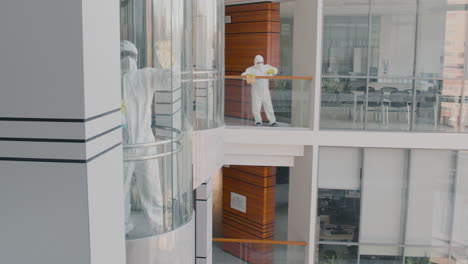 distant view of two cleaning men wearing personal protective equipment cleaning stair railings and glass elevator inside an office building