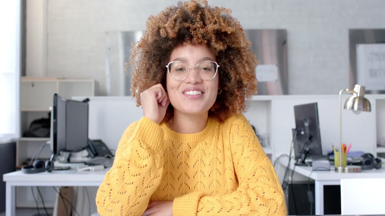 Felice donna d'affari di etnia mista in abiti casual con acconciatura afro e occhiali che effettua una videochiamata in ufficio, al rallentatore