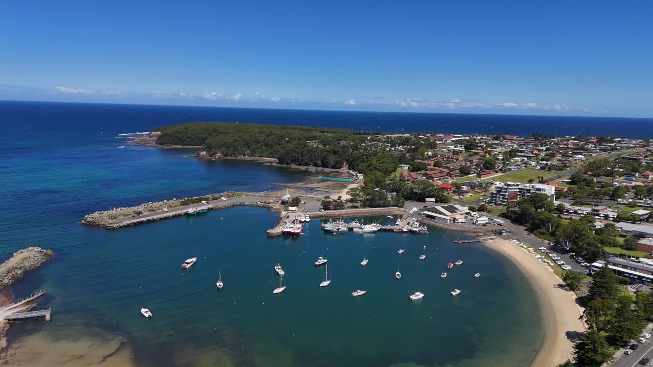 Aerial of Ulladulla Harbour with boats, calm blue water, and coastal town surrounds, establishing orbit
