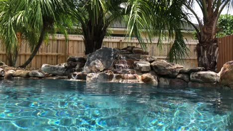 Peaceful-outdoor-pool-setting-with-palm-trees,-blue-salt-water-and-a-bubbling-waterfall