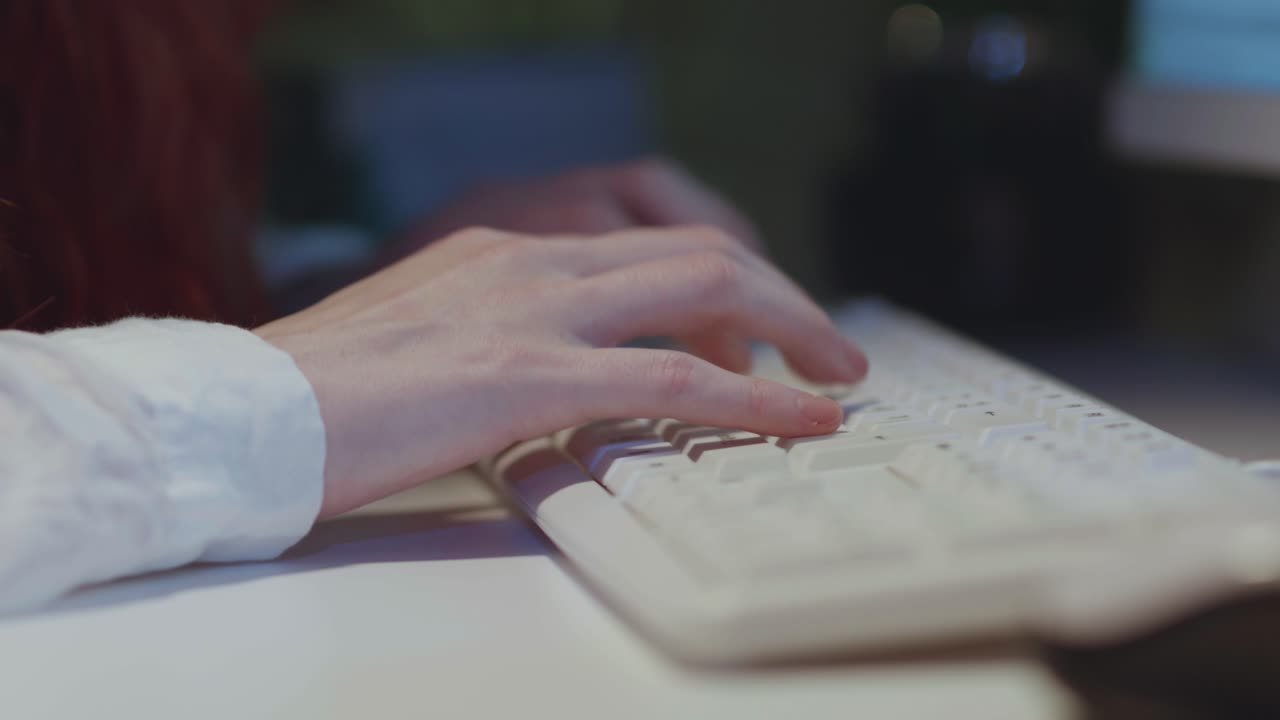 A close-up, side-angle shot of a woman's hands typing quickly on a white computer keyboard in a dimly lit office. This footage is for concepts of work, writing, coding, data entry, and communication.