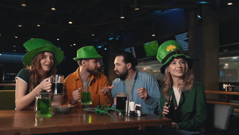 Friends watching a football game on TV in a pub, reacting excited and celebrating victory. Men and women having fun in Irish hats. Saint Patrick&#039;s Day.